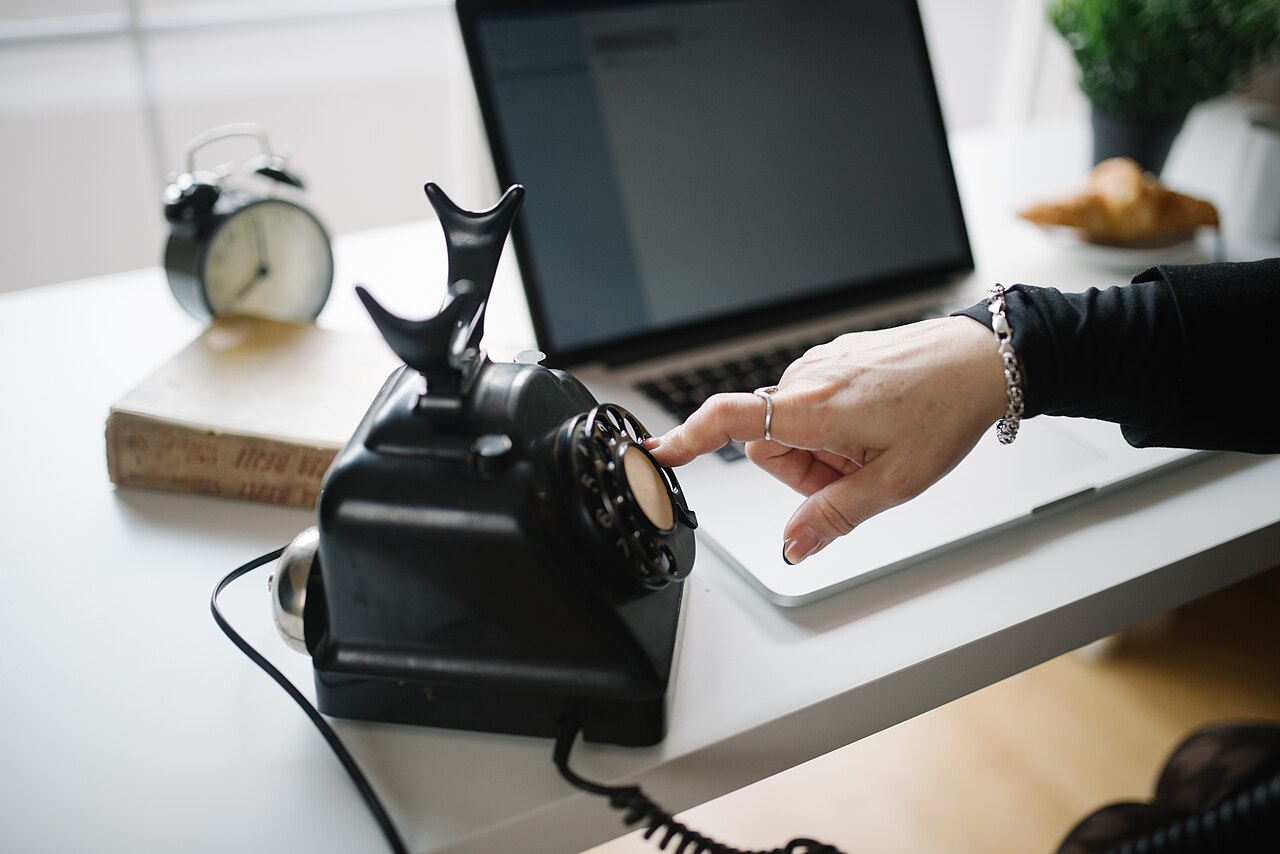A hand dialing a vintage rotary telephone on a desk next to a laptop
